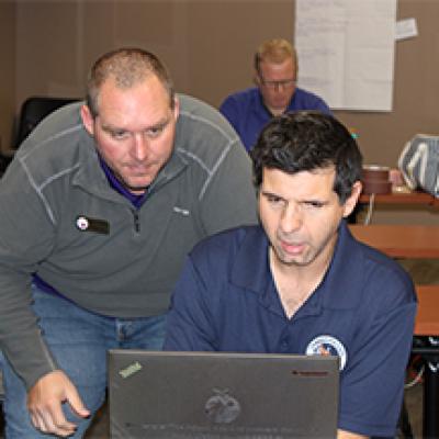 Man working at a laptop with another man looking over his shoulder offering assistance.