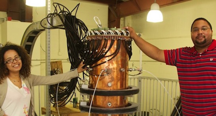 "Two people pose next to scientific equipment"
