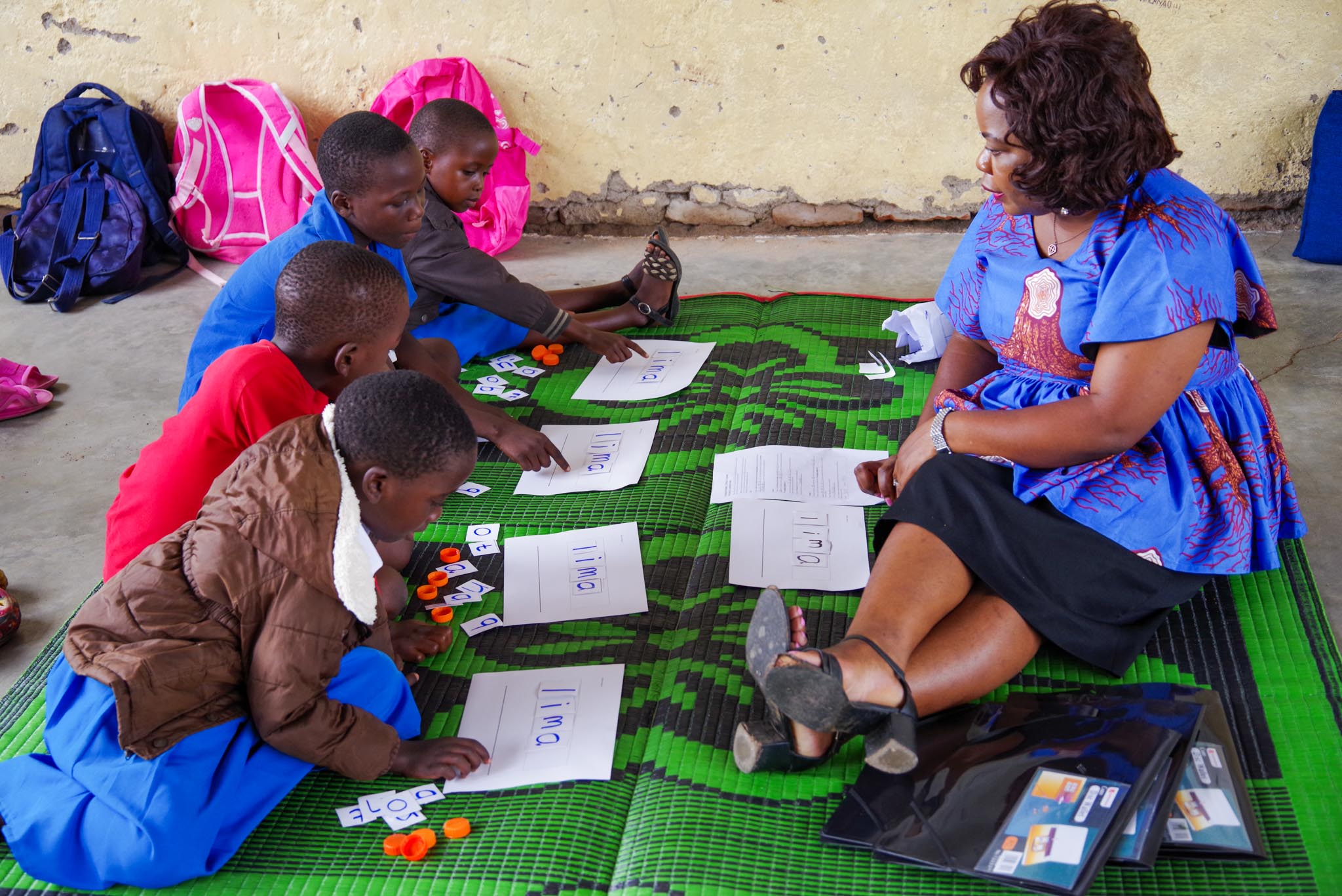 "A teacher is sitting on a carpet facing four young students who are all looking at a paper in front of them. "