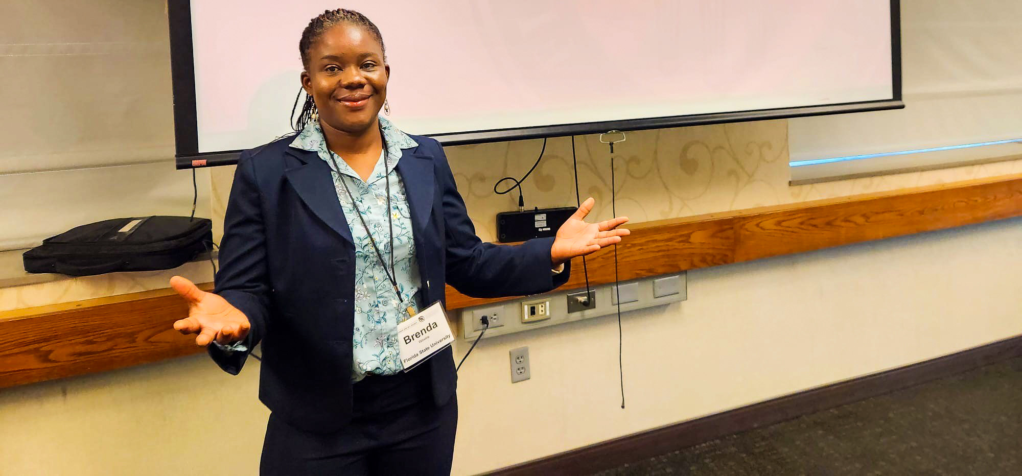 "Dr. Brenda Wawire at a conference posing in front of a screen showing her presentation title. "
