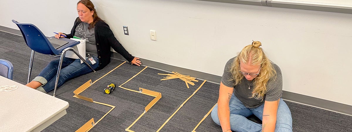 Two women sitting on the floor with laptops guiding a robot car through a maze.