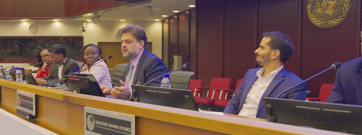 A panel discussion takes place in a conference room with several speakers seated behind a long desk equipped with microphones and monitors. One panelist speaks while others listen. Signage on the front of the desk reads “Educators Shaping Futures,” and the room features wood-paneled walls, overhead lighting, and rows of red chairs in the background.