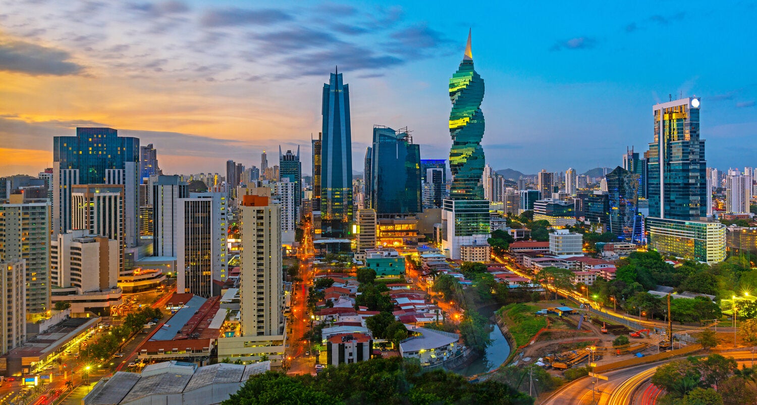 The image features a panoramic view of Panama City’s skyline at dusk, including modern high-rise buildings and illuminated city streets.