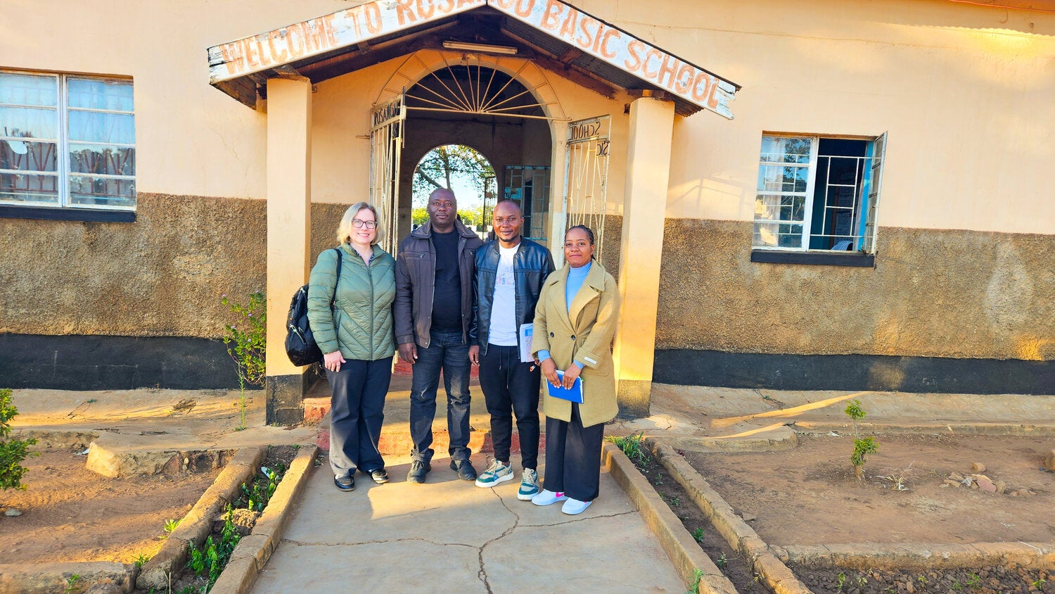 Four people stand outside a school building in Zambia, posing for a group photo.