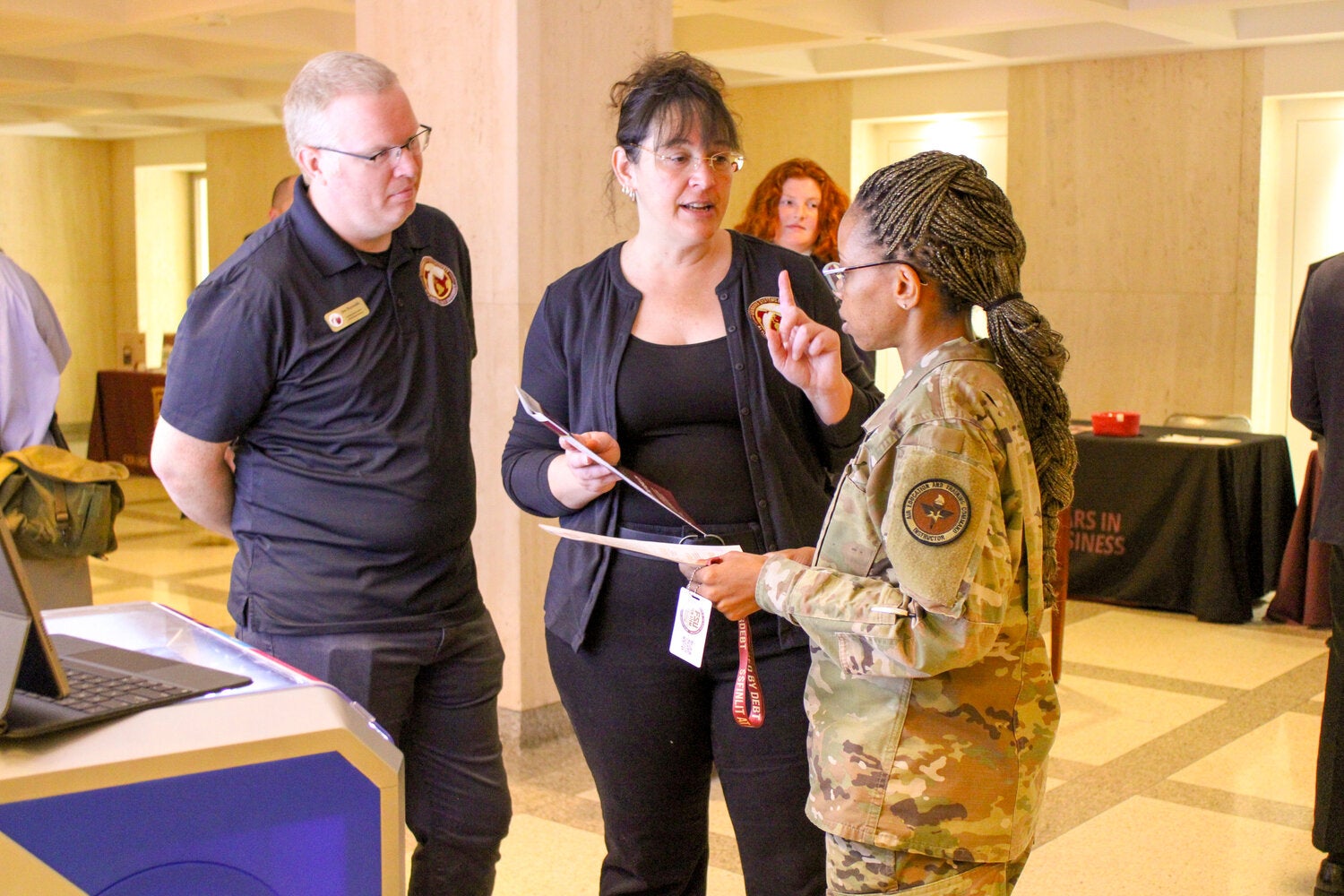 Three attendees converse beside an information table in a large lobby or conference space. One person in a military uniform holds documents while speaking with two individuals wearing matching logoed shirts and name badges. A laptop sits on the table in the foreground, and additional attendees and event displays are visible in the background.