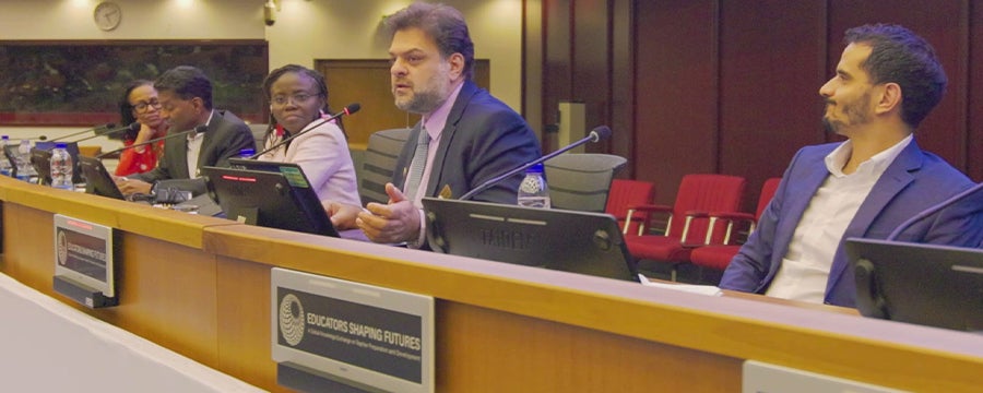 A panel discussion takes place in a conference room with several speakers seated behind a long desk equipped with microphones and monitors. One panelist speaks while others listen. Signage on the front of the desk reads “Educators Shaping Futures,” and the room features wood-paneled walls, overhead lighting, and rows of red chairs in the background.