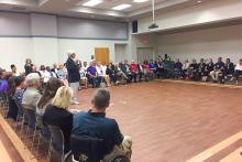 A large group sitting in chairs around the perimeter of the room listening to a man talking with a microphone