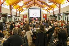 Wide shot of a full banquet hall with a speaker at the front under a large projection screen.