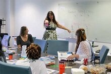 Carrie Meyers standing at a white board pointing towards participants around a table listening to her presentation.