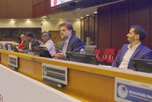 A panel discussion takes place in a conference room with several speakers seated behind a long desk equipped with microphones and monitors. One panelist speaks while others listen. Signage on the front of the desk reads “Educators Shaping Futures,” and the room features wood-paneled walls, overhead lighting, and rows of red chairs in the background.