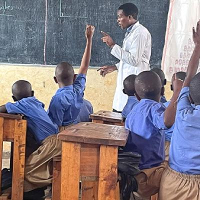 A classroom of students in Rwanda with hands raised and a teacher in front of the room by a chalkboard.