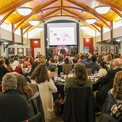 Wide shot of a full banquet hall with a speaker at the front under a large projection screen.