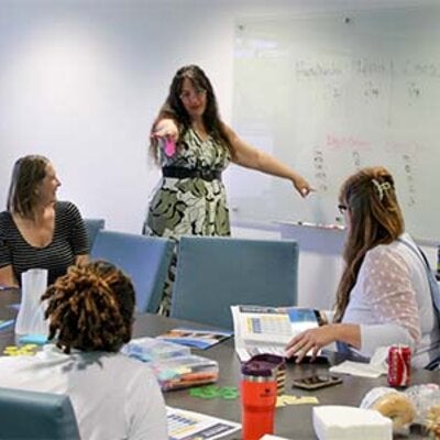 Carrie Meyers standing at a white board pointing towards participants around a table listening to her presentation.