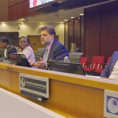 A panel discussion takes place in a conference room with several speakers seated behind a long desk equipped with microphones and monitors. One panelist speaks while others listen. Signage on the front of the desk reads “Educators Shaping Futures,” and the room features wood-paneled walls, overhead lighting, and rows of red chairs in the background.