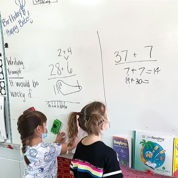 "Two children standing at a whiteboard working on math problems. "