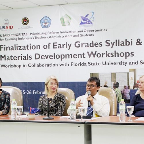 "Group sitting at a dais. There is a man holding a microphone. The banner behind them says Finalization of Early Grade Syllabi and Materials Development Workshop"