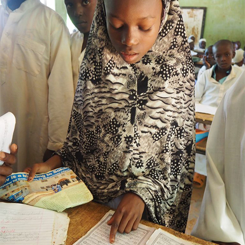"A young girl in a Northern Nigerian classroom standing in front of a book and pointing to a section"
