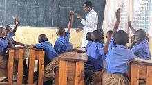 A classroom of students in Rwanda with hands raised and a teacher in front of the room by a chalkboard.