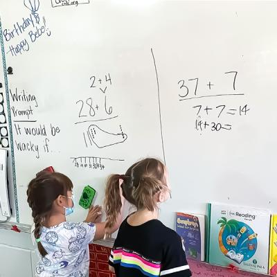 Two children standing at a whiteboard working on math problems. 